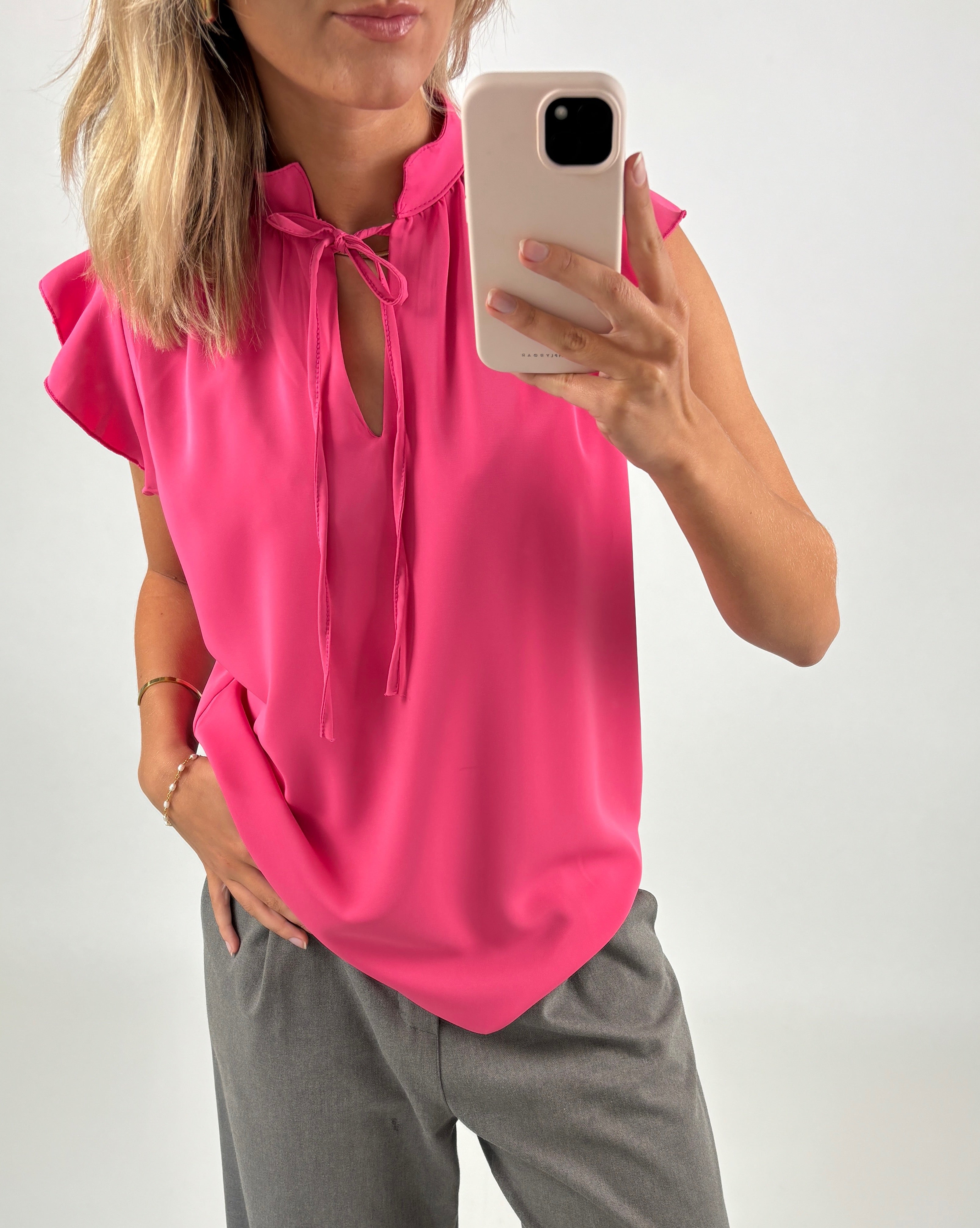 En person iført en pink bluse tager et billede med en smartphone. Personen står oprejst og befinder sig i et neutralt, lys miljø.