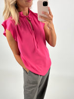 En person tager et spejlfoto iført en pink bluse og grå bukser i et lyst rum. Personen holder en mobiltelefon.