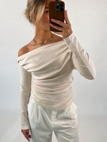 Person tager selfie iført cremefarvet off-shoulder bluse og hvide bukser, står i en neutral, lys baggrund.