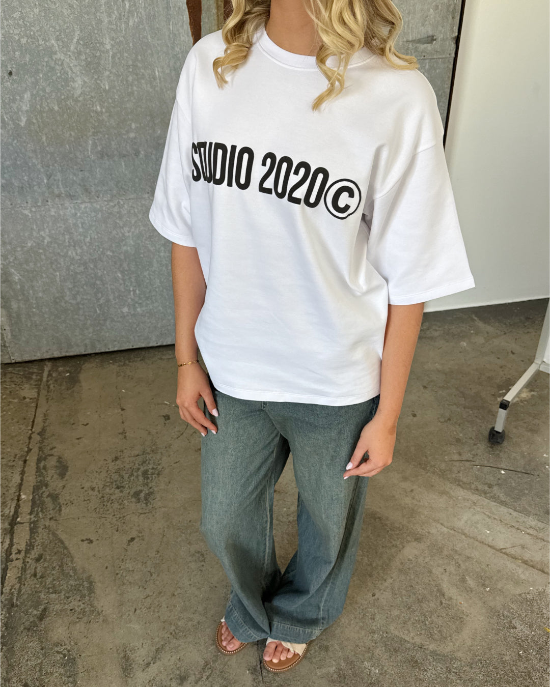 En person står iført en hvid t-shirt med teksten "STUDIO 2020©," grønne bukser og sandaler, på et betonunderlag foran en grå væg.