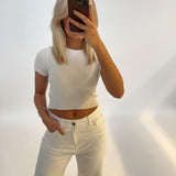 En person iført en hvid t-shirt tager et spejlbillede med en smartphone i et lyst rum, der har en ren, ensfarvet baggrund.