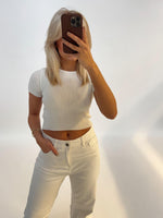 En person iført en hvid t-shirt tager et spejlbillede med en smartphone i et lyst rum, der har en ren, ensfarvet baggrund.