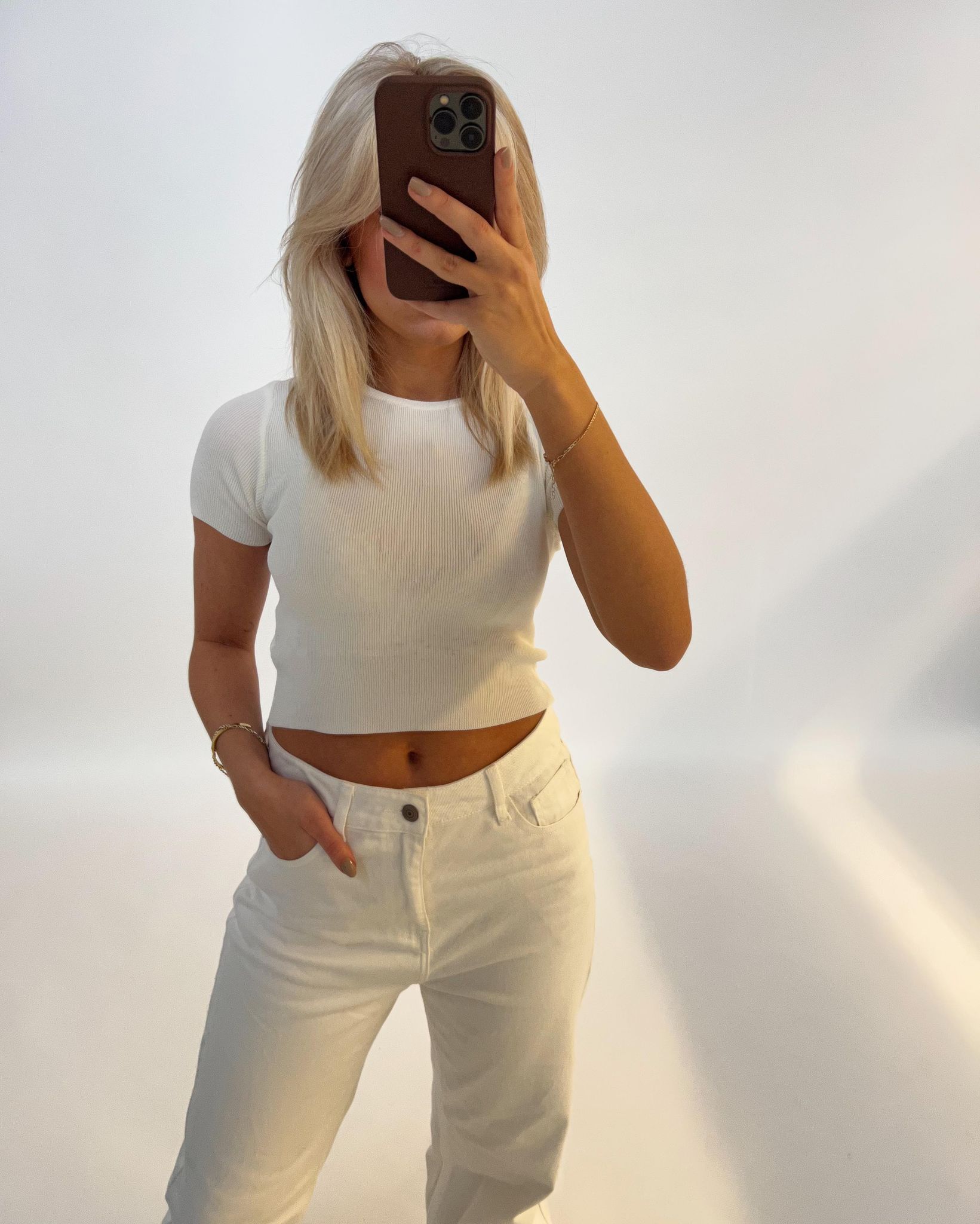 En person iført en hvid t-shirt tager et spejlbillede med en smartphone i et lyst rum, der har en ren, ensfarvet baggrund.