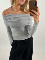 En person iført en grå off-shoulder bluse, tager en selfie med en smartphone, mens hun står i et rum med lys baggrund. Hun har rød neglelak og bærer sort bukser.