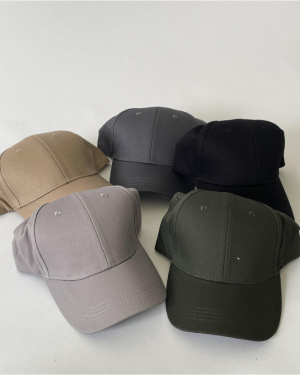 Fem baseballkasketter ligger på en hvid overflade. Kasketterne er i forskellige farver: beige, grå, sort, lysegrå og olivengrøn, arrangeret i en halvcirkel.