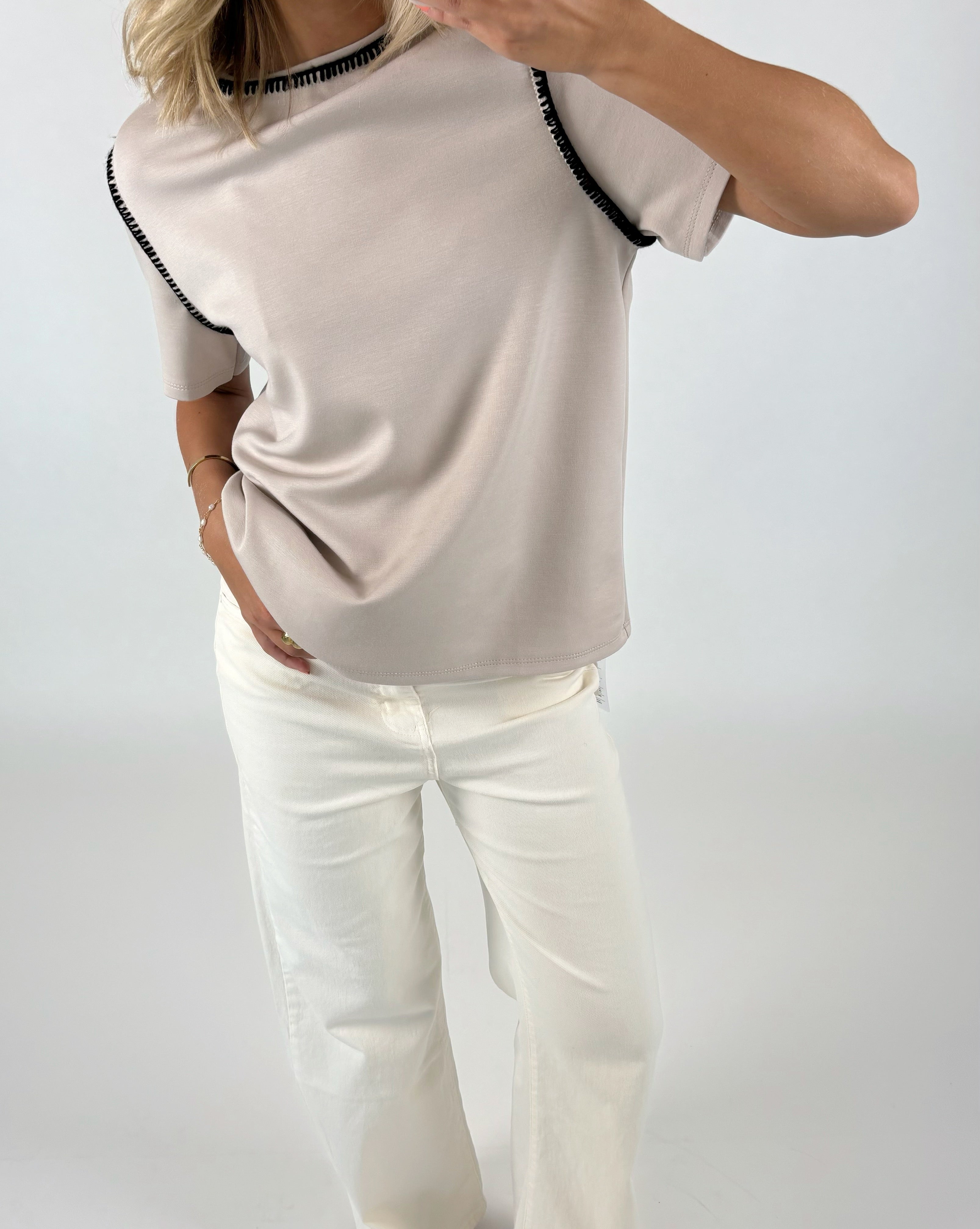 Person iført lys beige t-shirt med sorte kanter, poserer med en hånd i lommen på hvide bukser. Baggrunden er en lys, ensfarvet overflade.