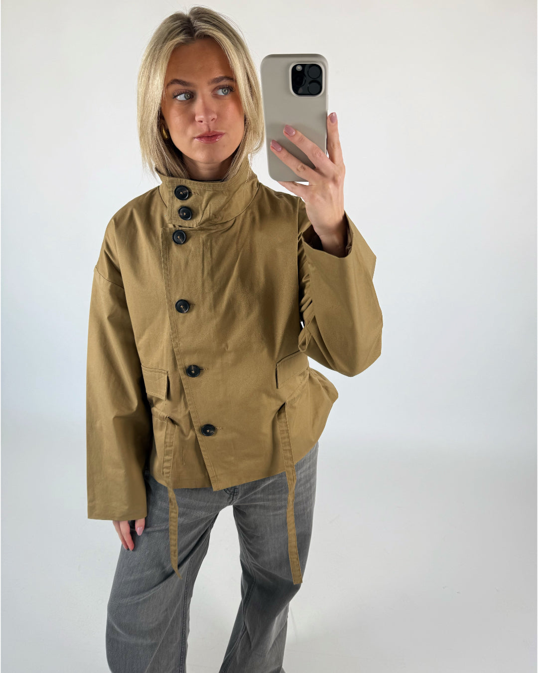 Person tager selfie, iført brun jakke og grå bukser, i et neutralt rum. Hun holder en telefon med et lyst cover og kigger væk fra kameraet.