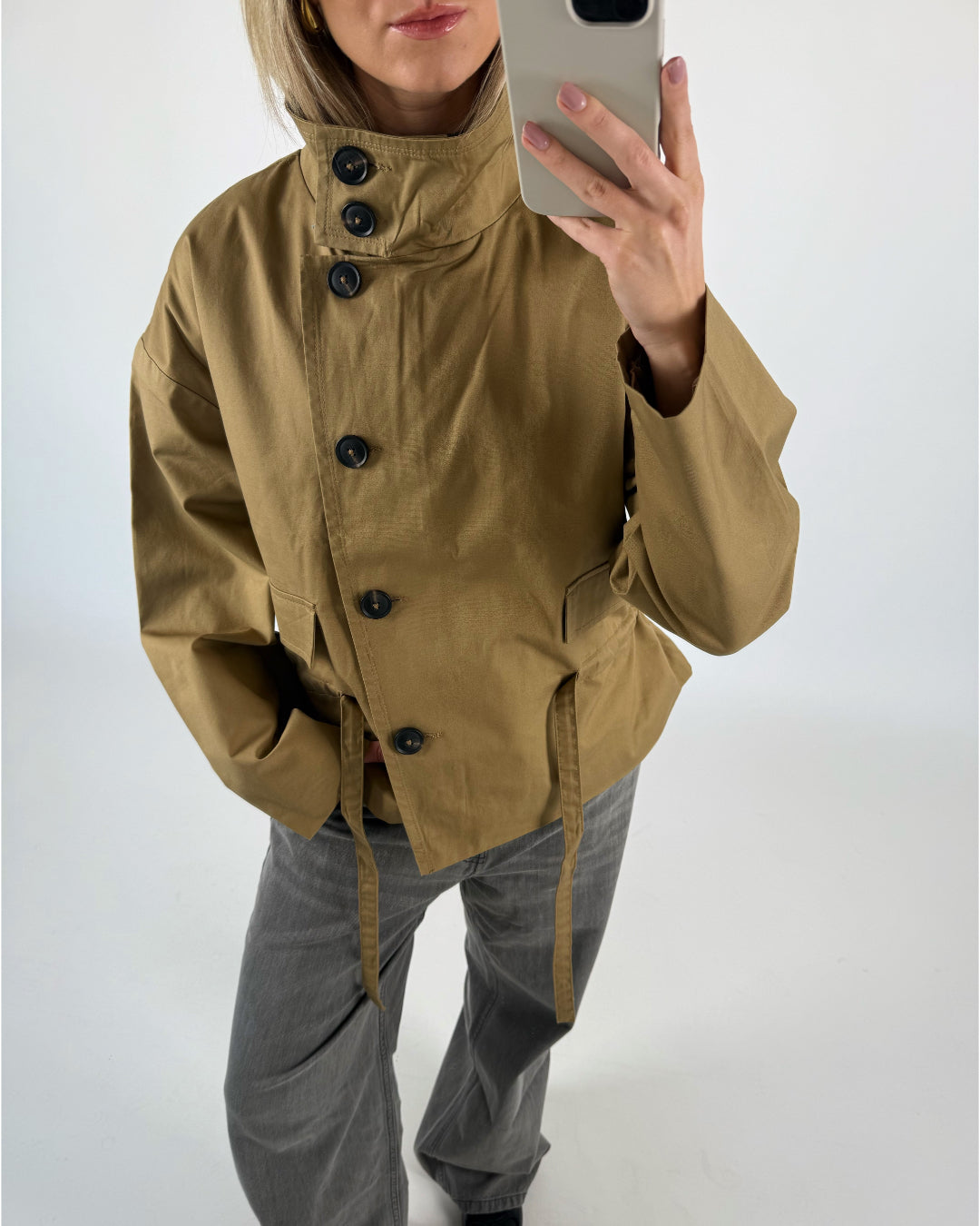 En person iført brun jakke med store knapper fotograferer sig selv med en mobiltelefon. Baggrunden er enkel og grå, hvilket fremhæver personens outfit i billedet.