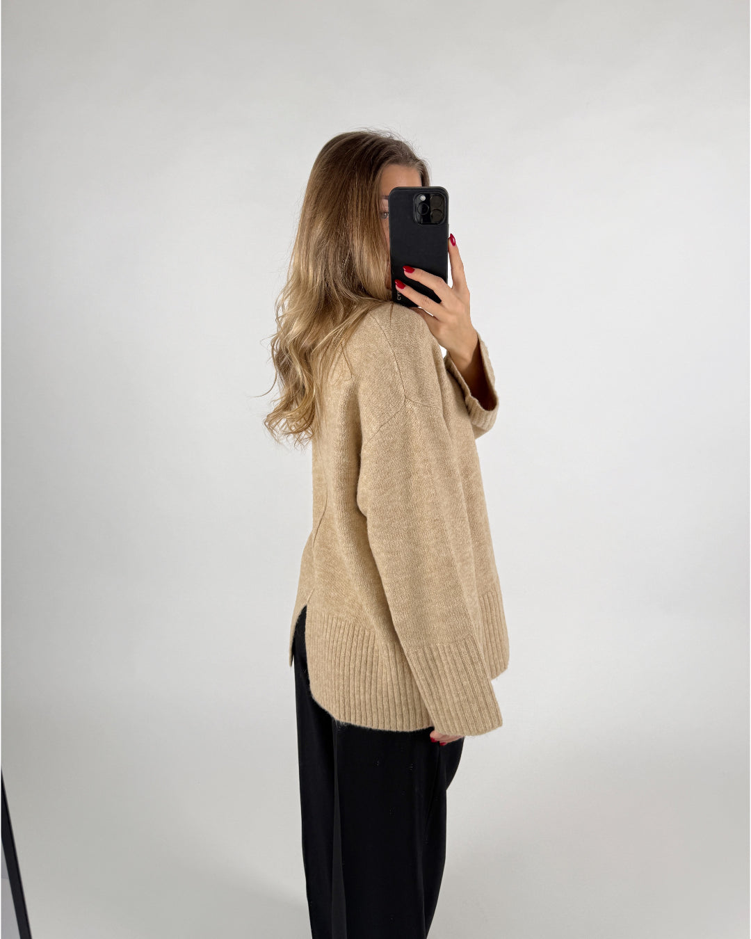 Kvinde iført beige sweater og sorte bukser, tager et spejlbillede med sin telefon. Hun står i en lys, minimalistisk indretning.
