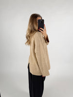 Kvinde iført beige sweater og sorte bukser, tager et spejlbillede med sin telefon. Hun står i en lys, minimalistisk indretning.