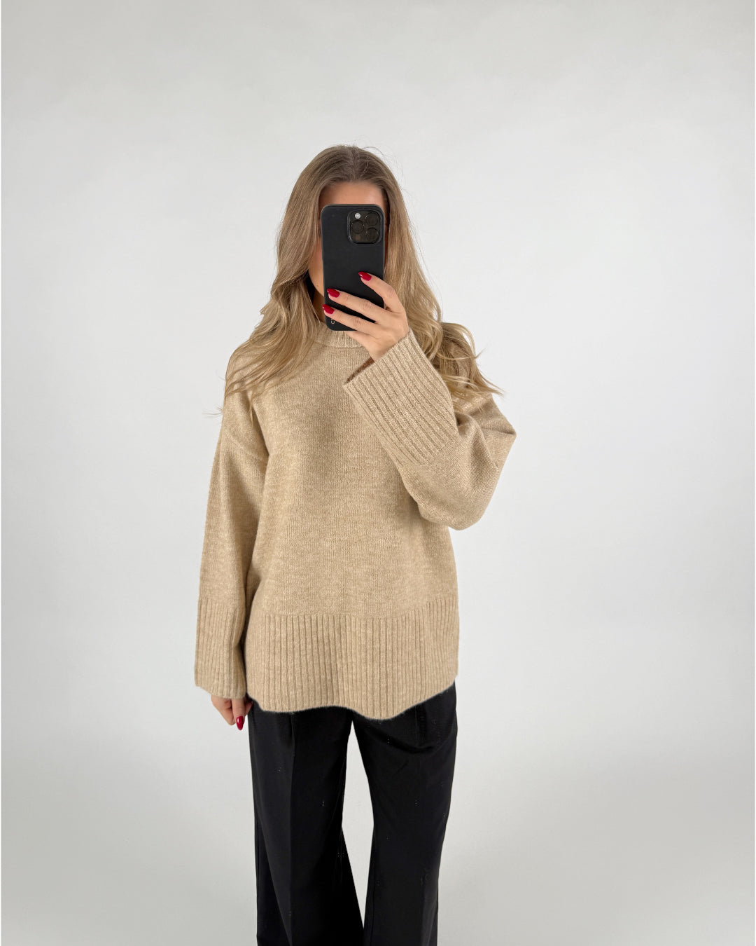 En person iført en beige sweater tager et selfie med en mobiltelefon, mens de står foran en lys baggrund. De har løst, bølget hår og sorte bukser.