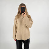 En person iført en beige sweater tager et selfie med en mobiltelefon, mens de står foran en lys baggrund. De har løst, bølget hår og sorte bukser.