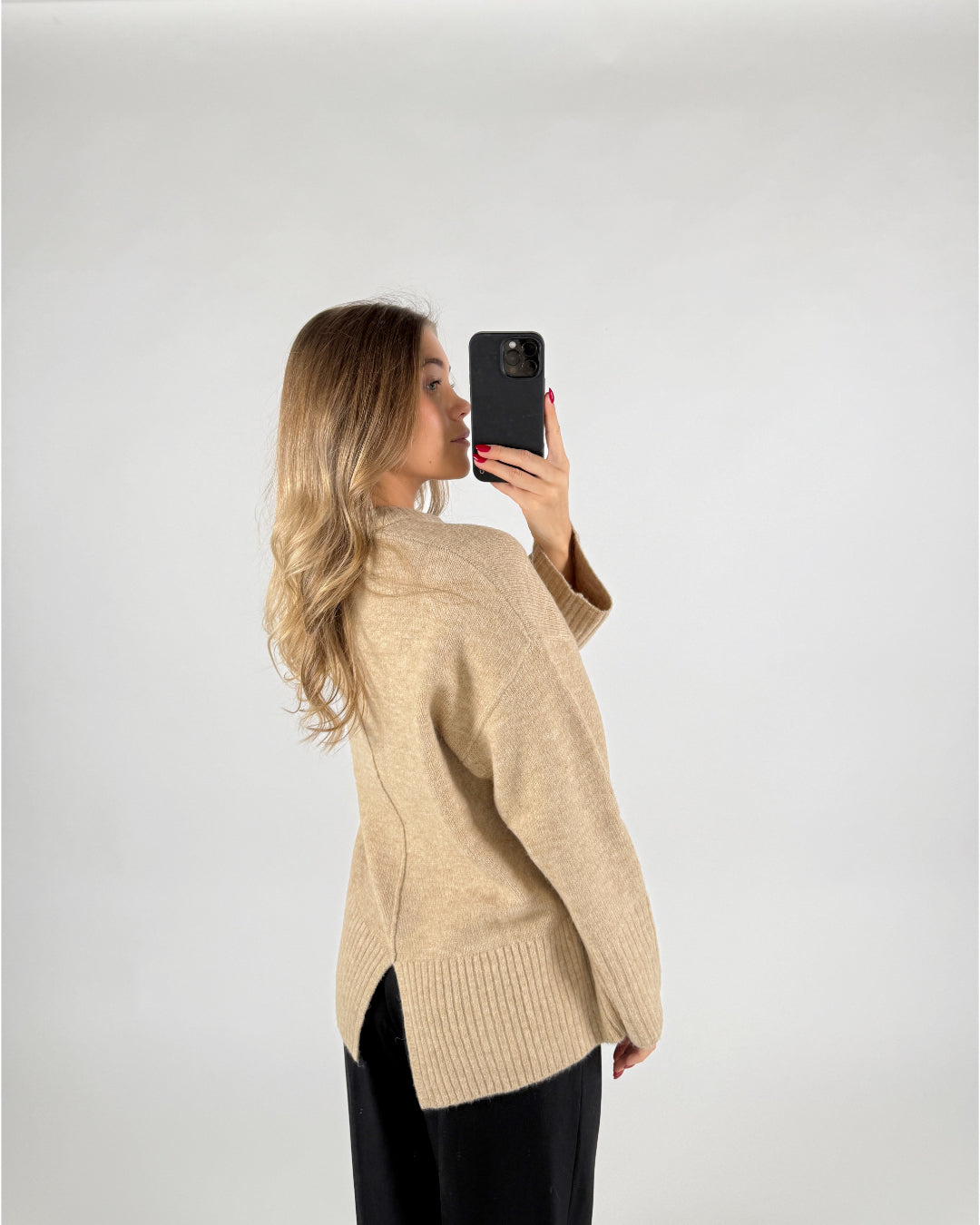 En kvinde i en beige sweater tager et selfie med sin telefon mod en lys baggrund. Hun har langt, bølget hår og bærer sorte bukser. Ingen tekst er til stede.