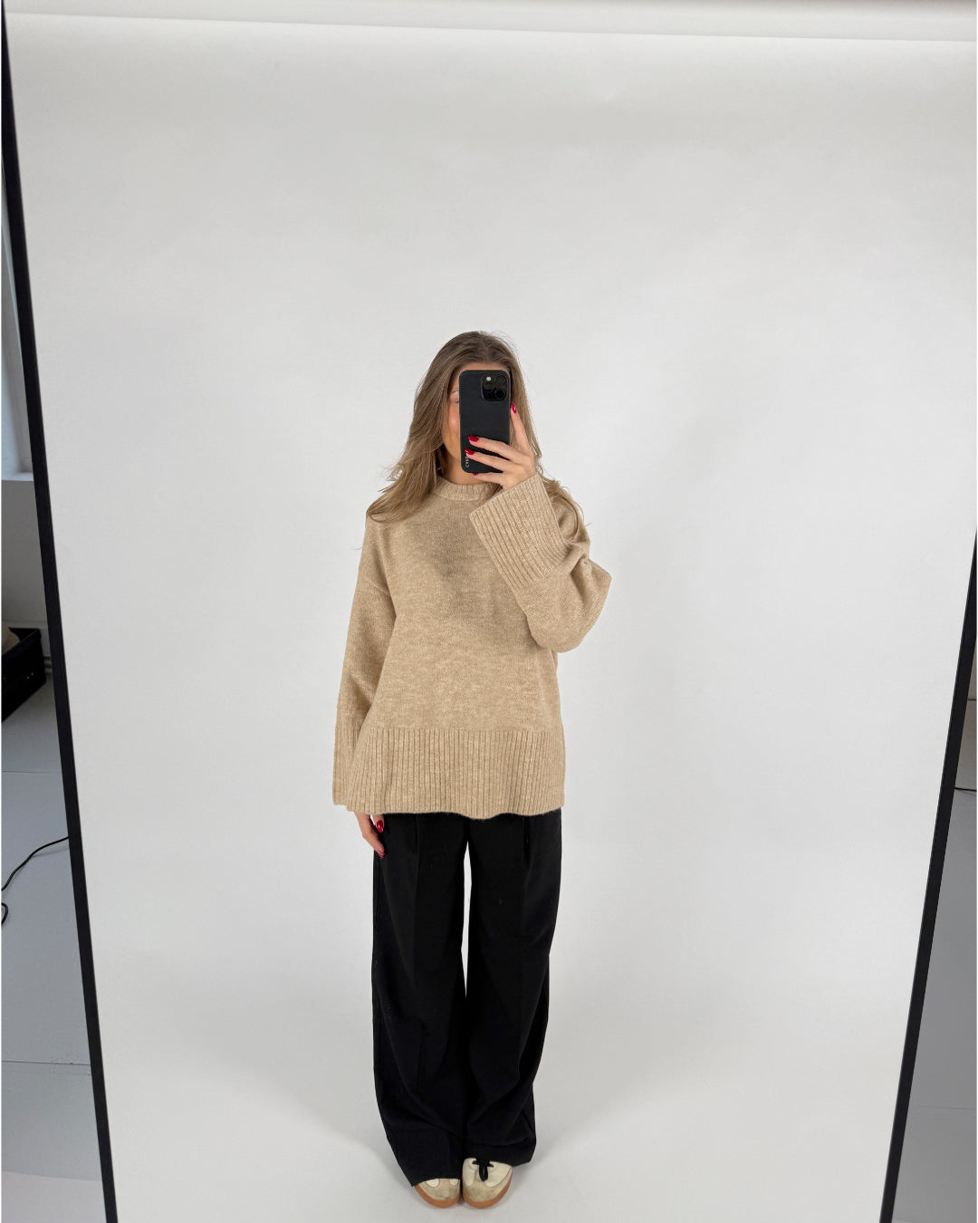 En person står i et lyst rum og tager et spejlbillede iført en beige sweater og sorte bukser. Telefonen dækker ansigtet.