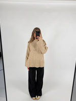 En person står i et lyst rum og tager et spejlbillede iført en beige sweater og sorte bukser. Telefonen dækker ansigtet.