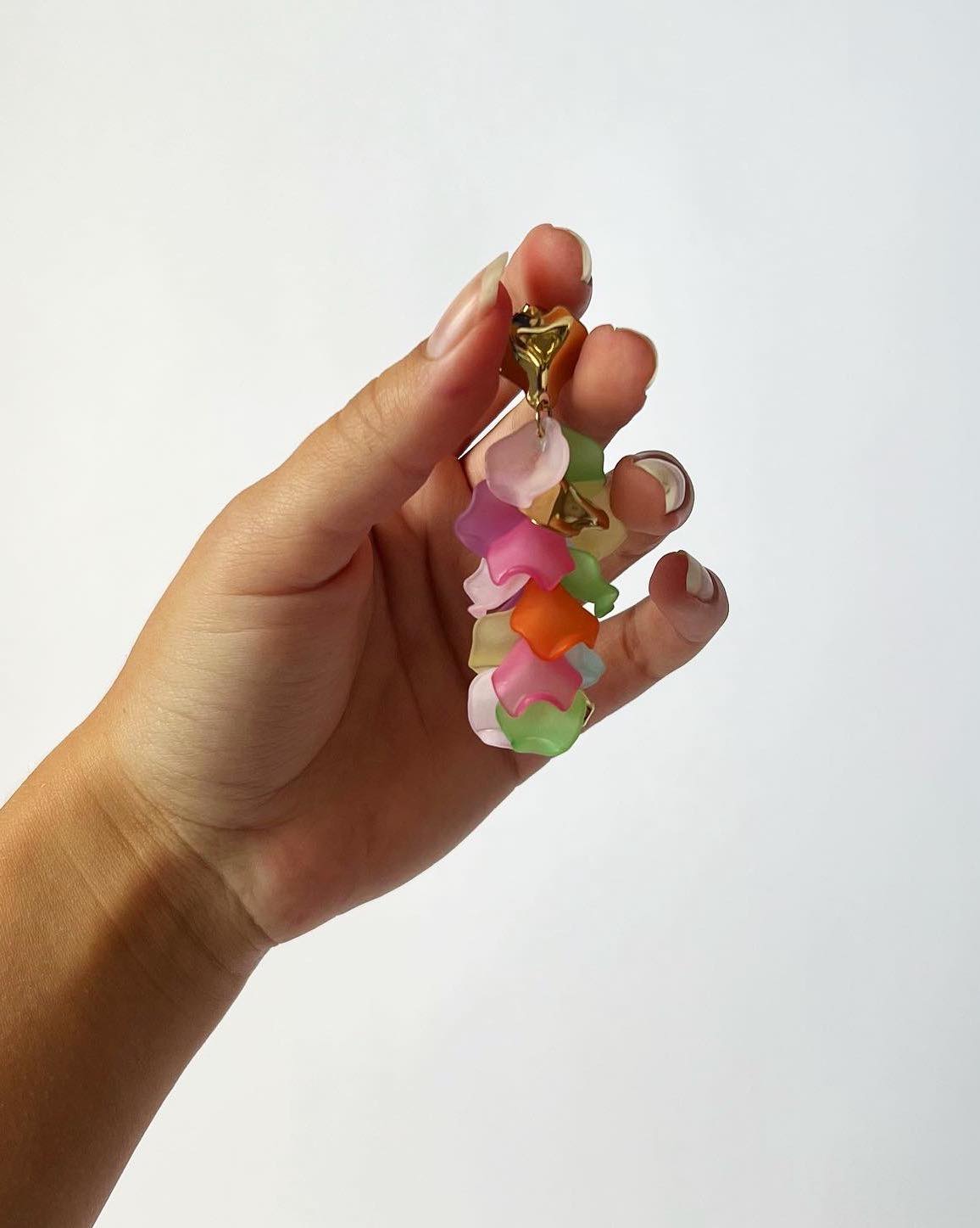 Hånden holder et farverigt nøgleringvedhæng med flere lag af blomstformede plaststykker i pink, orange, grøn og gul toner på en hvid baggrund.