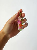 Hånden holder et farverigt nøgleringvedhæng med flere lag af blomstformede plaststykker i pink, orange, grøn og gul toner på en hvid baggrund.