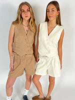 To kvinder iført matchende vest-og-shorts-sæt, den ene i beige, den anden i hvid, poserer sammen mod en hvid baggrund, iført afslappet fodtøj og smykker.
