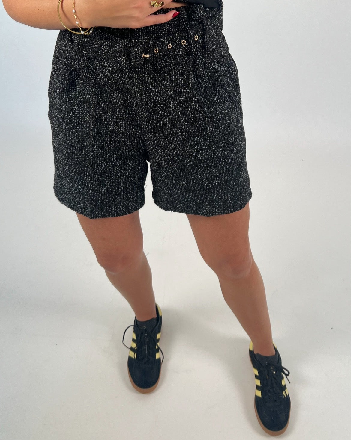 En person iført sorte shorts med mønster står med en hånd ved taljen. De bærer også sorte og gule sneakers på en hvid baggrund.