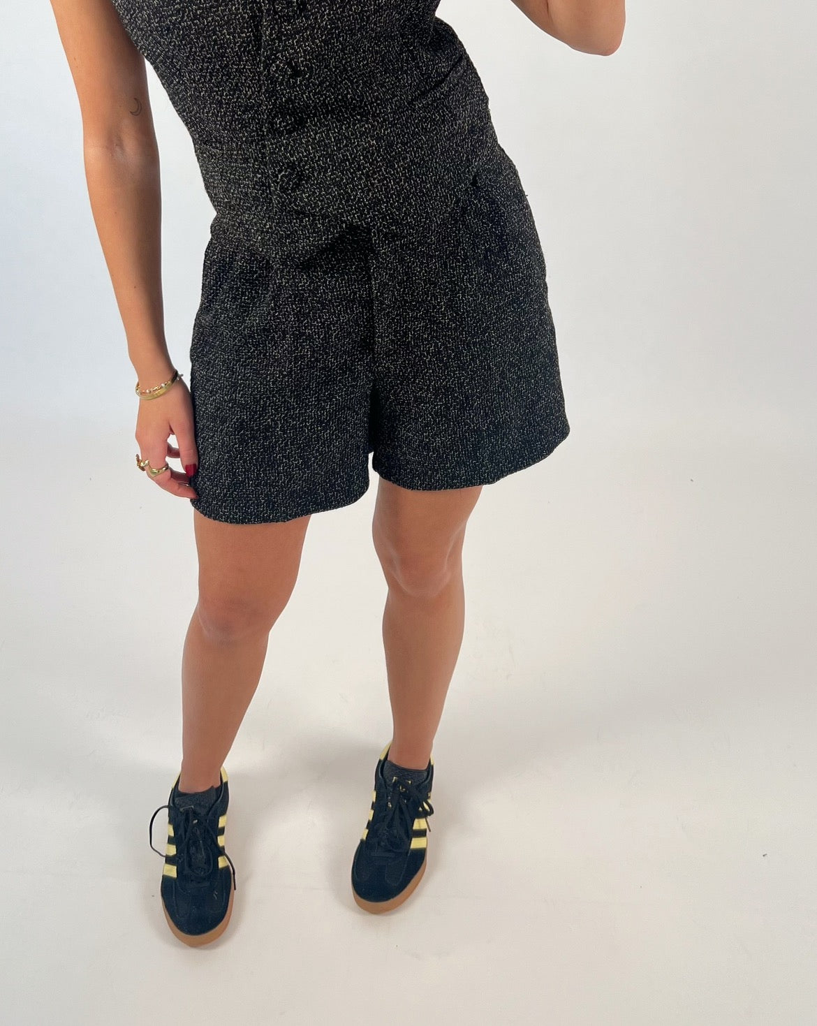 Person iført sort mønstret shorts og top, står på et hvidt gulv. De bærer sorte sneakers med gule striber og guldsmykker på håndleddet.