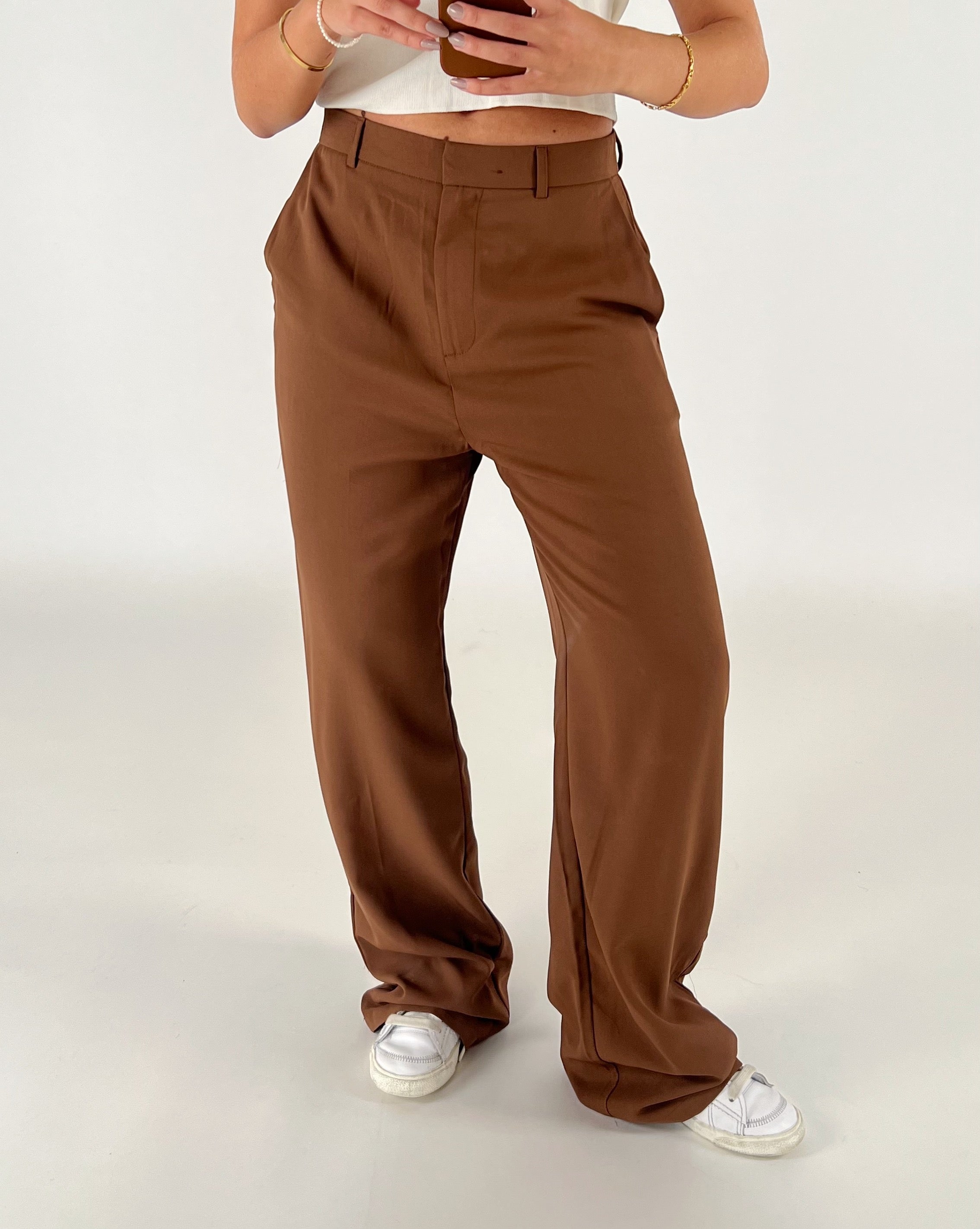 Brune bukser bæres, holdes oppe af en person i en hvid top og hvide sneakers, mens personen holder en telefon. Baggrunden er enkel og lys.