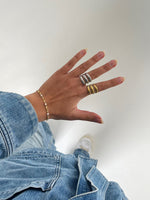 En hånd bærer to flettede ringe og et armbånd. Personen iført blå denimjakke og jeans står på en lys baggrund.
