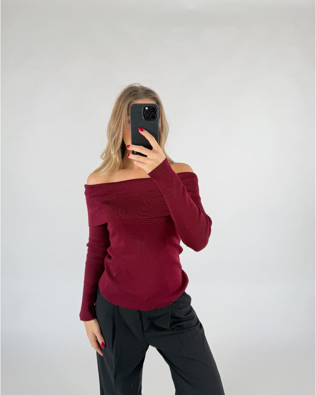 En person iført en mørkerød off-shoulder trøje tager et spejlbillede med en sort telefon i et minimalistisk rum med lysegrå baggrund.