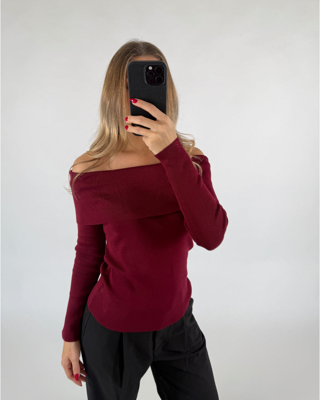 En person iført en rød trøje holder en sort smartphone op foran ansigtet og tager et billede. Baggrunden er lys og enkel.