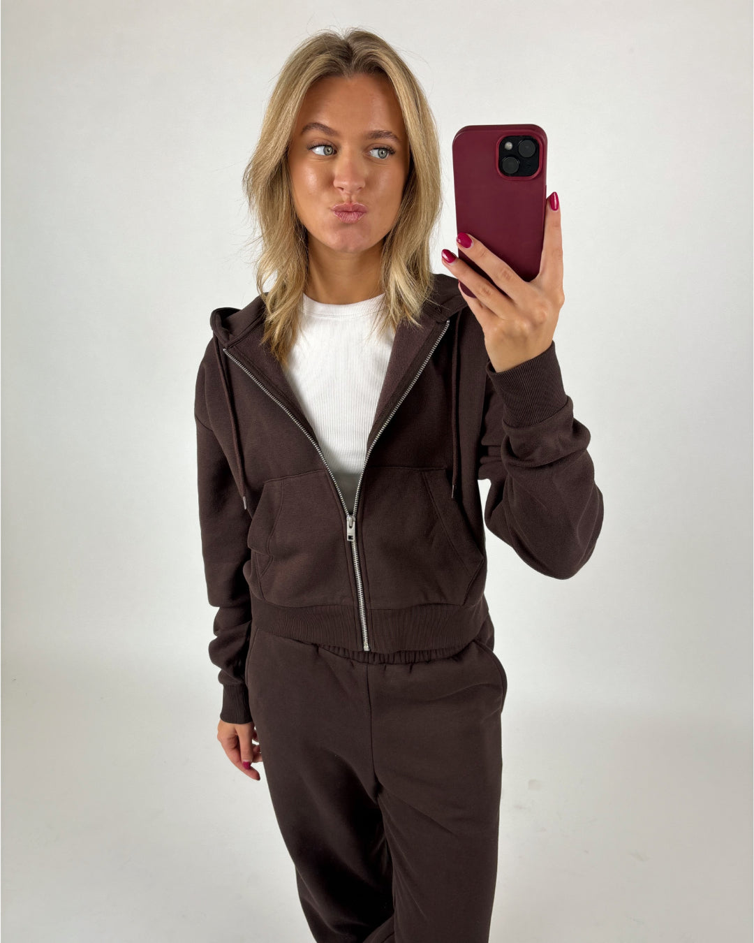 Kvinde tager selfie iført brun hættetrøje og bukser, stående foran en lys baggrund. Hun laver en sjov ansigtsudtryk, mens hun holder sin telefon med et mørkerødt cover.