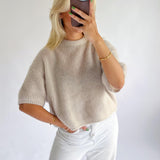 En person tager en selfie med en smartphone. De har en lys beige striktrøje og hvide bukser på. Personen har grønlakerede negle og bærer guldsmykker, stående mod en hvid baggrund.