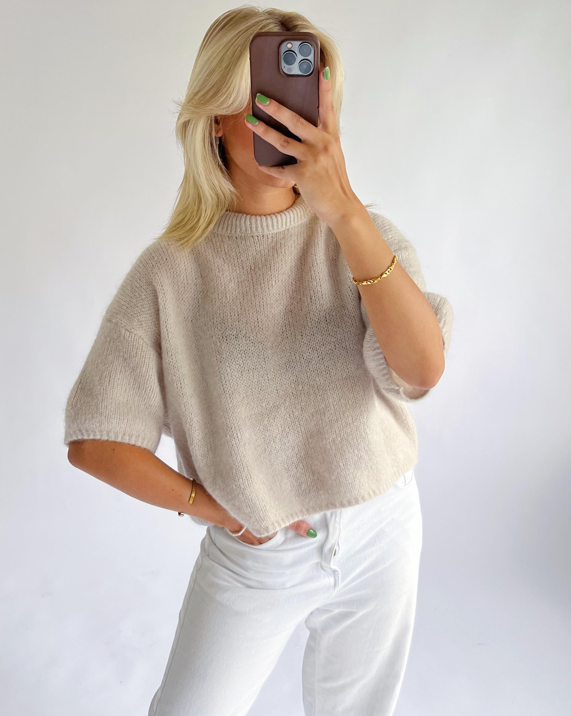 En person tager en selfie med en smartphone. De har en lys beige striktrøje og hvide bukser på. Personen har grønlakerede negle og bærer guldsmykker, stående mod en hvid baggrund.