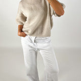 En person iført en løs, beige sweater putter hånden i lommen på hvide bukser. Personen står oprejst i et neutralt, lys rum.