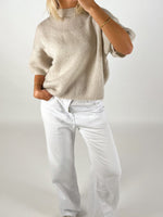 En person iført en løs, beige sweater putter hånden i lommen på hvide bukser. Personen står oprejst i et neutralt, lys rum.