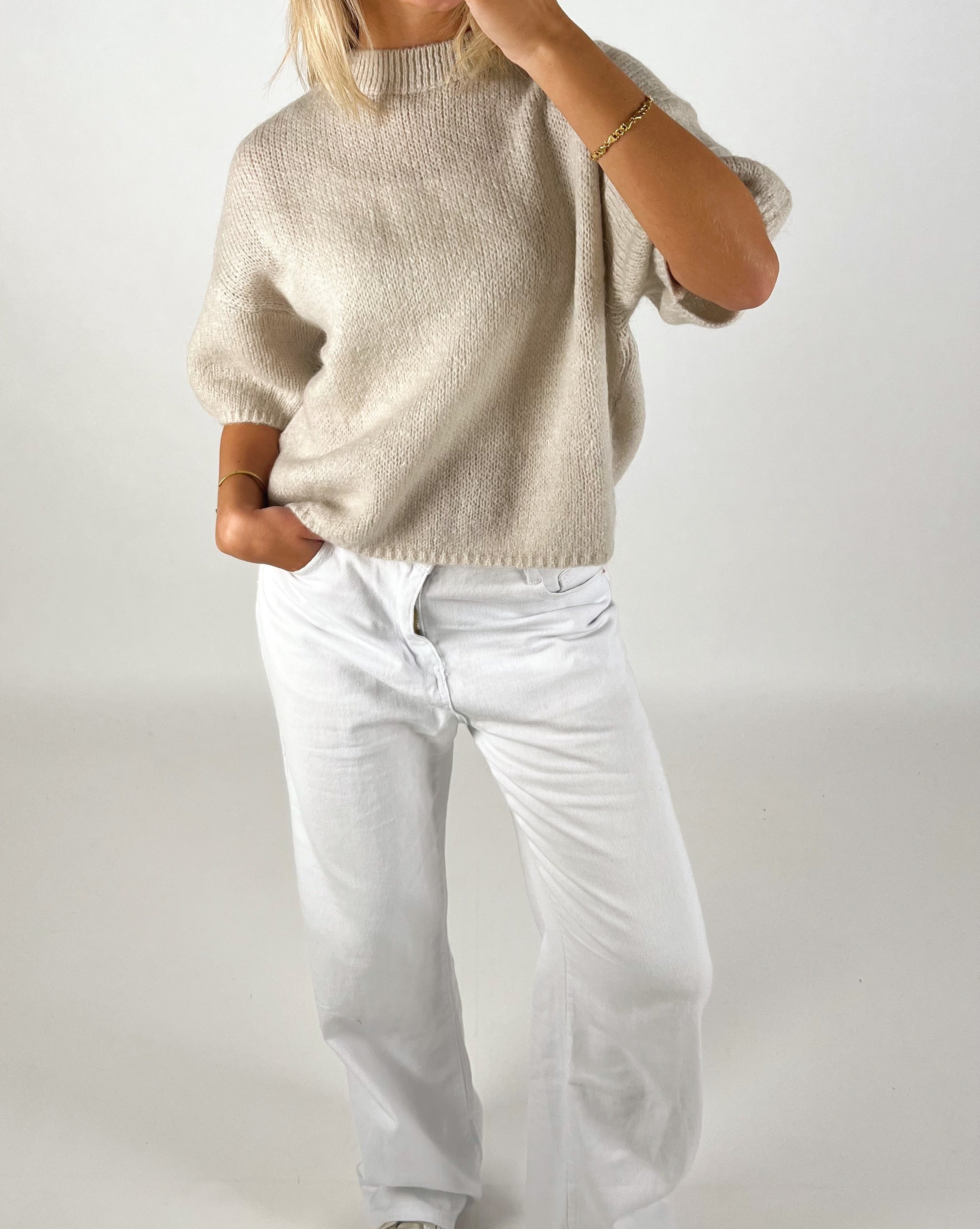 En person iført en løs, beige sweater putter hånden i lommen på hvide bukser. Personen står oprejst i et neutralt, lys rum.
