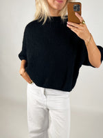 En person iført en sort sweater og hvide bukser tager et spejlbillede med en smartphone i et enkelt, lyst rum.