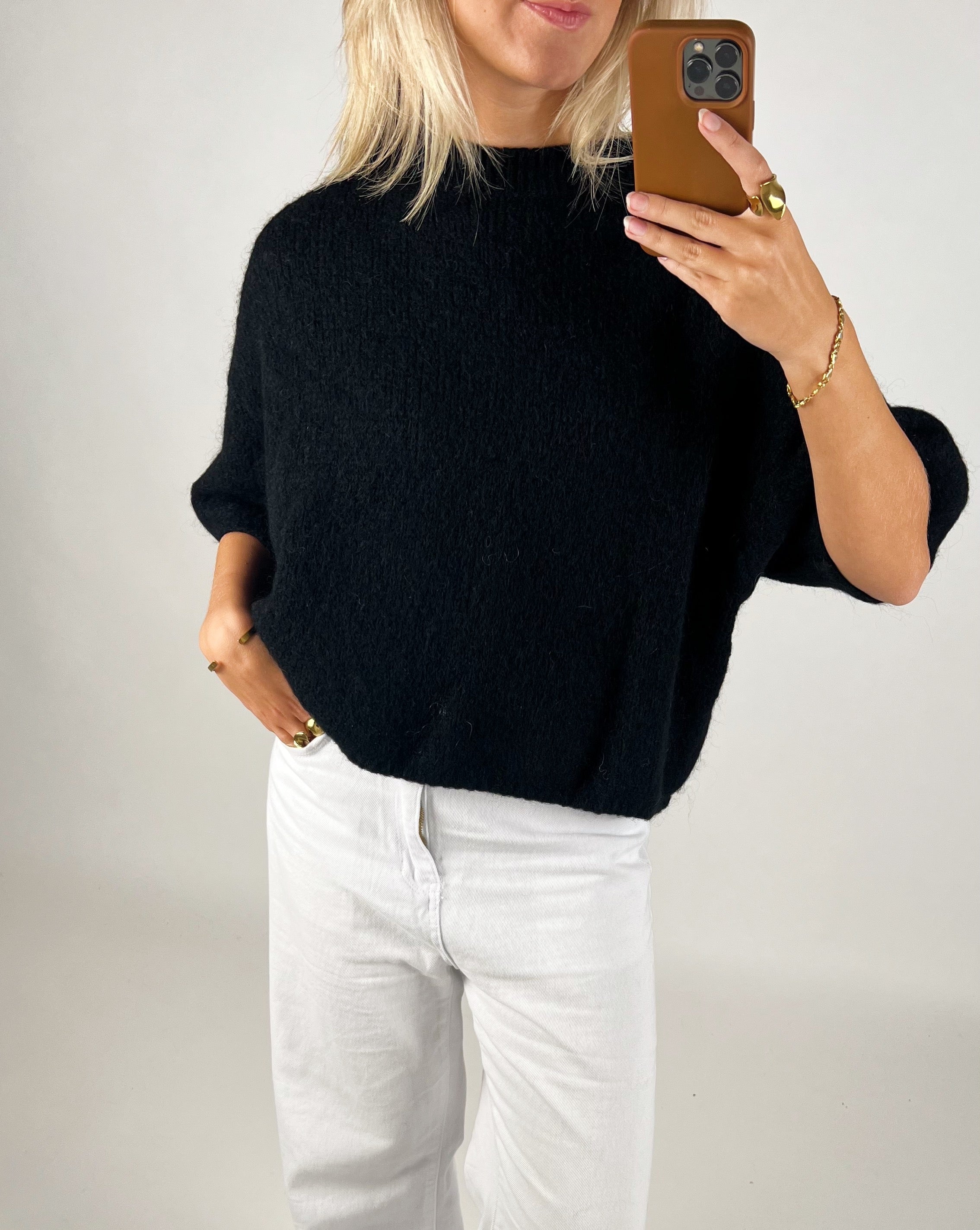 En person iført en sort sweater og hvide bukser tager et spejlbillede med en smartphone i et enkelt, lyst rum.