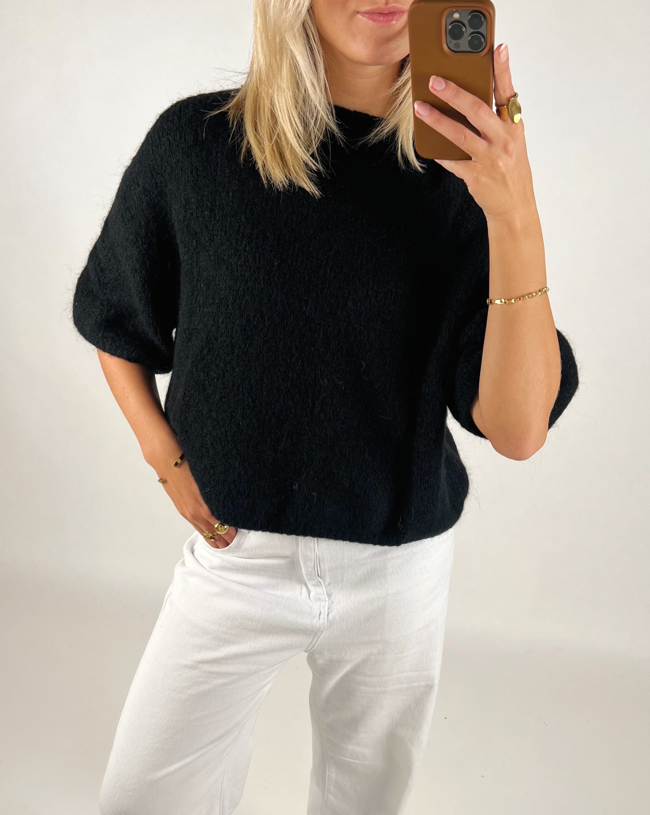 En person i sort trøje og hvide bukser tager et spejlbillede med en mobiltelefon. Personen har lyshår og enkle smykker. Baggrunden er ensfarvet hvid.