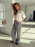 En person står afslappet med hånden i håret, iført en hvid sweater og grå jeans, i et lyst studie med vinduer, fotograferingsudstyr og moderne indretning.