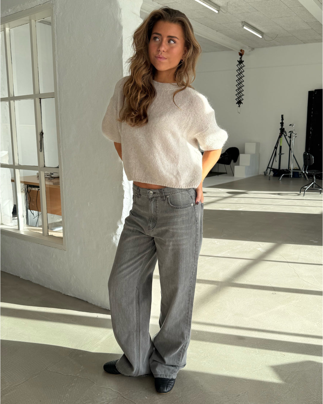En person iført hvid sweater og grå jeans står afslappet i et lyst studie med fotoudstyr og møbler i baggrunden.