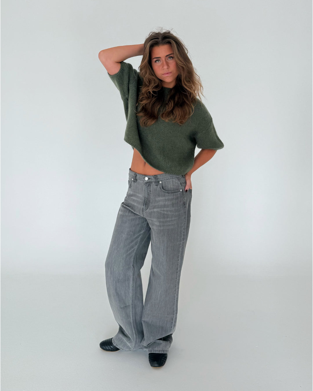En person iført en grøn sweater og grå jeans står med en hånd på hovedet i et enkelt, hvidt rum.