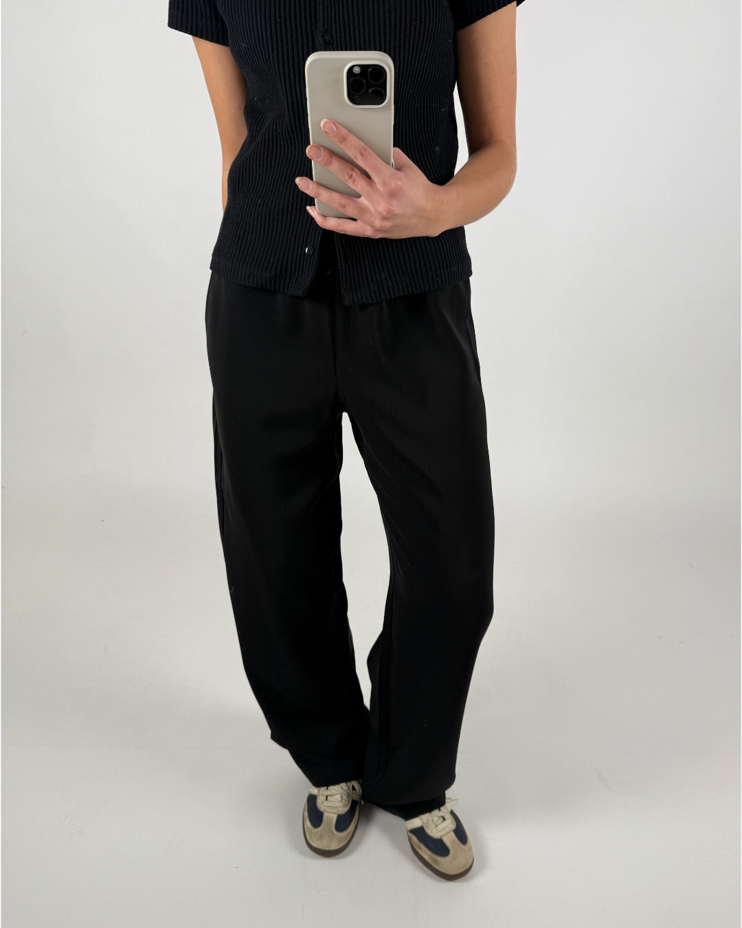 En person i sort tøj holder en smartphone i en lys baggrund. Personen har en mørk overdel, vide bukser og beige-sorte sneakers. Det er en minimalistisk indstilling.