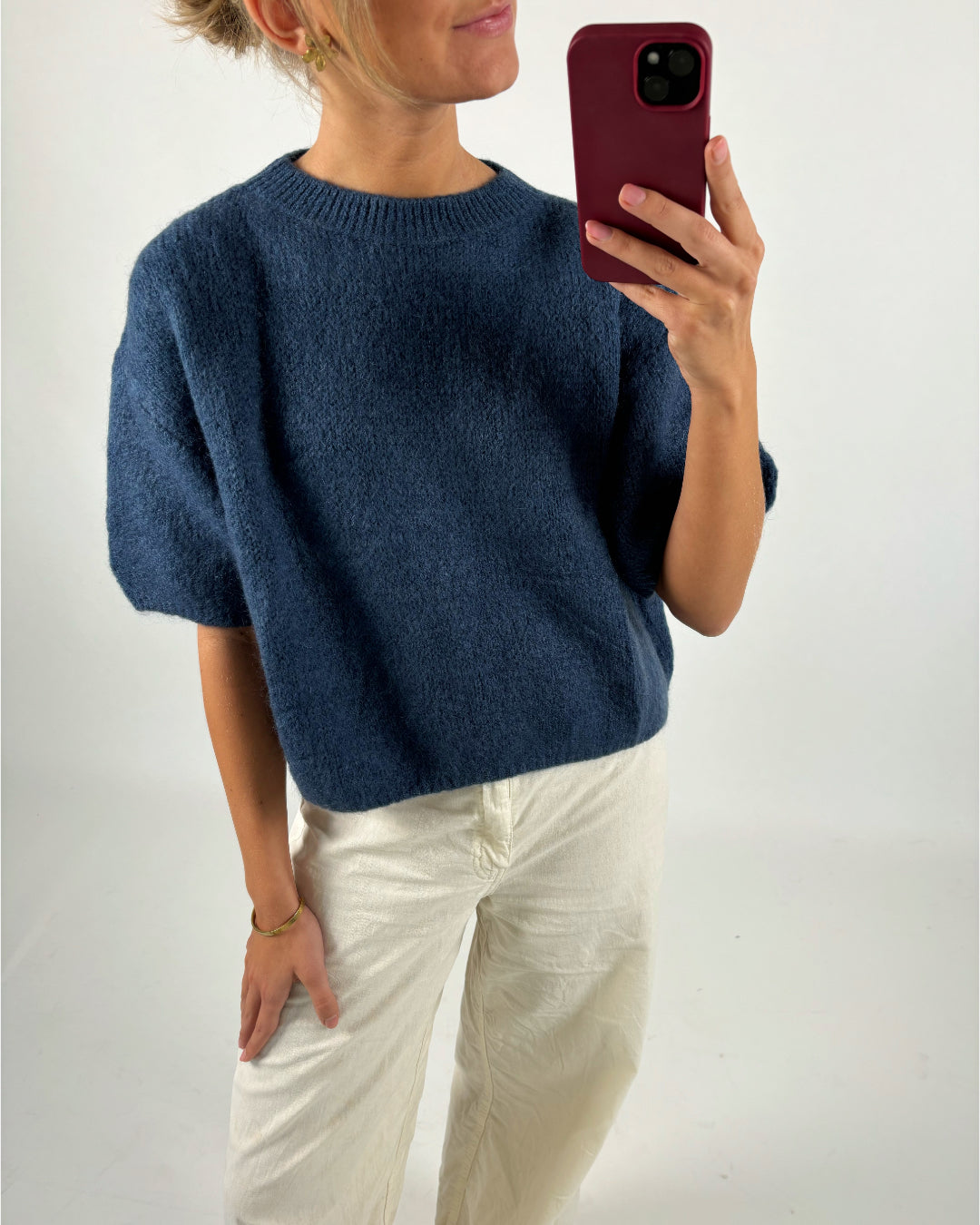 Personen bærer en blå striktrøje og hvide bukser, mens de tager en selfie med en telefon i rød etui, i et neutralt, oplyst rum.