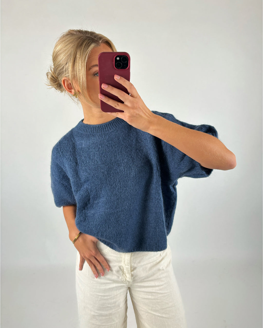 En person tager en selfie med en smartphone, iført en blå striktrøje og cremefarvede bukser, i et neutralt rum.
