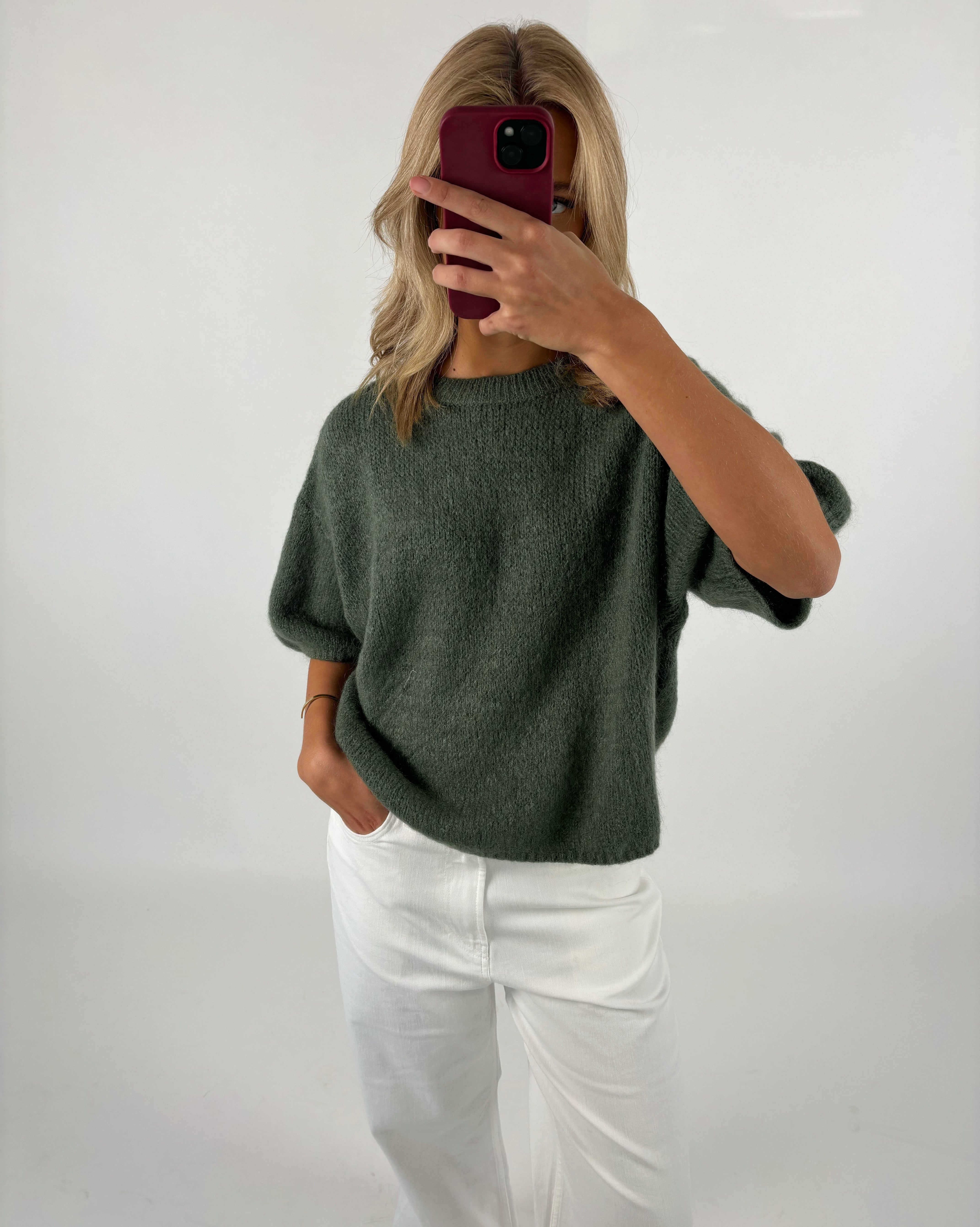 Person tager et spejlbillede med en smartphone, klædt i en grøn sweater og hvide bukser, mod en simpel, hvid baggrund.