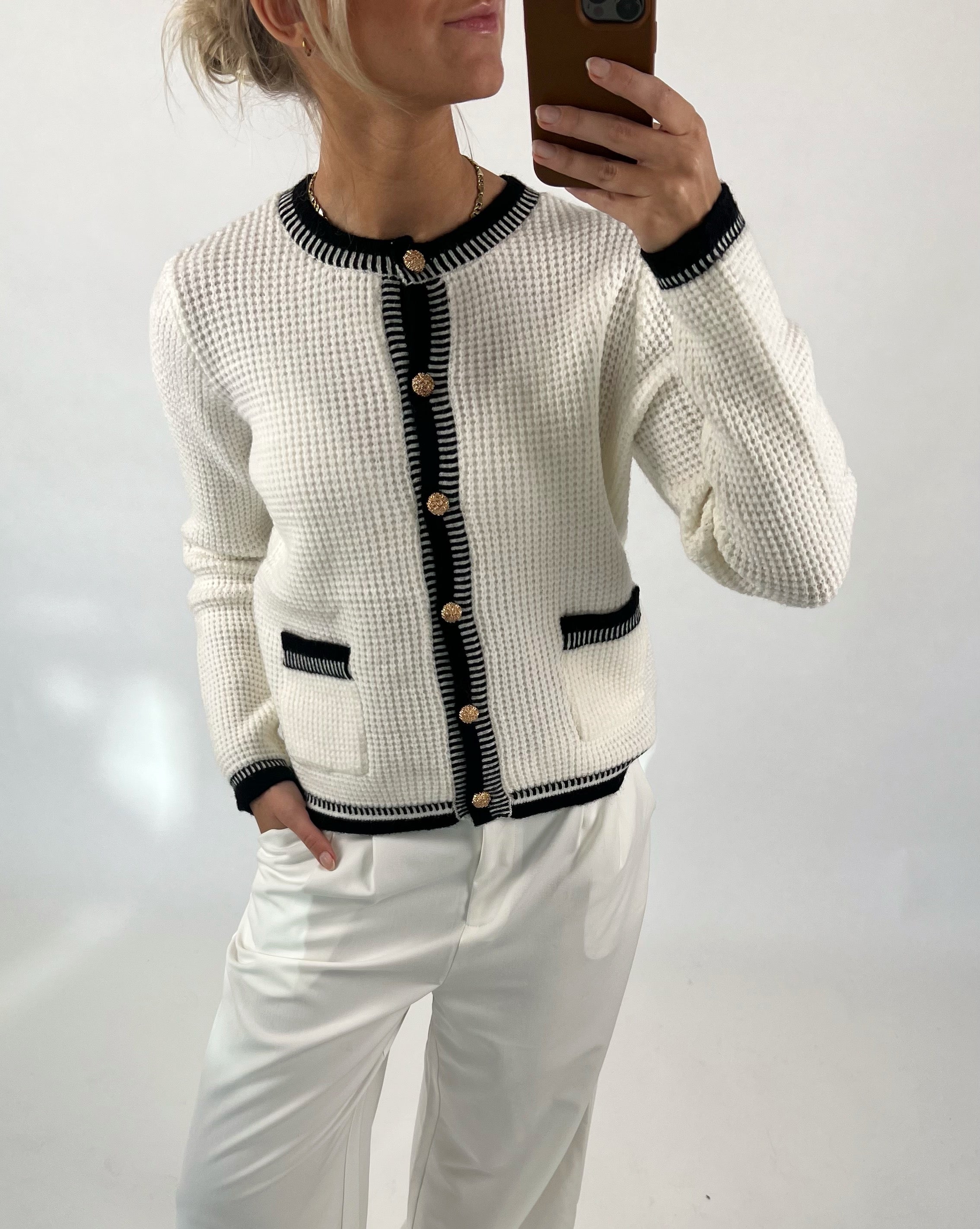 En person tager en selfie iført en hvid cardigan med sorte kanter og guldknapper. Personen står op mod en hvid baggrund og bærer hvide bukser.