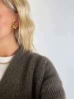 En person med lyst hår bærer en guldfarvet blomsterørering. Personen er iført en brun striktrøje og en hvid bluse med halskæde, mod en neutral baggrund.