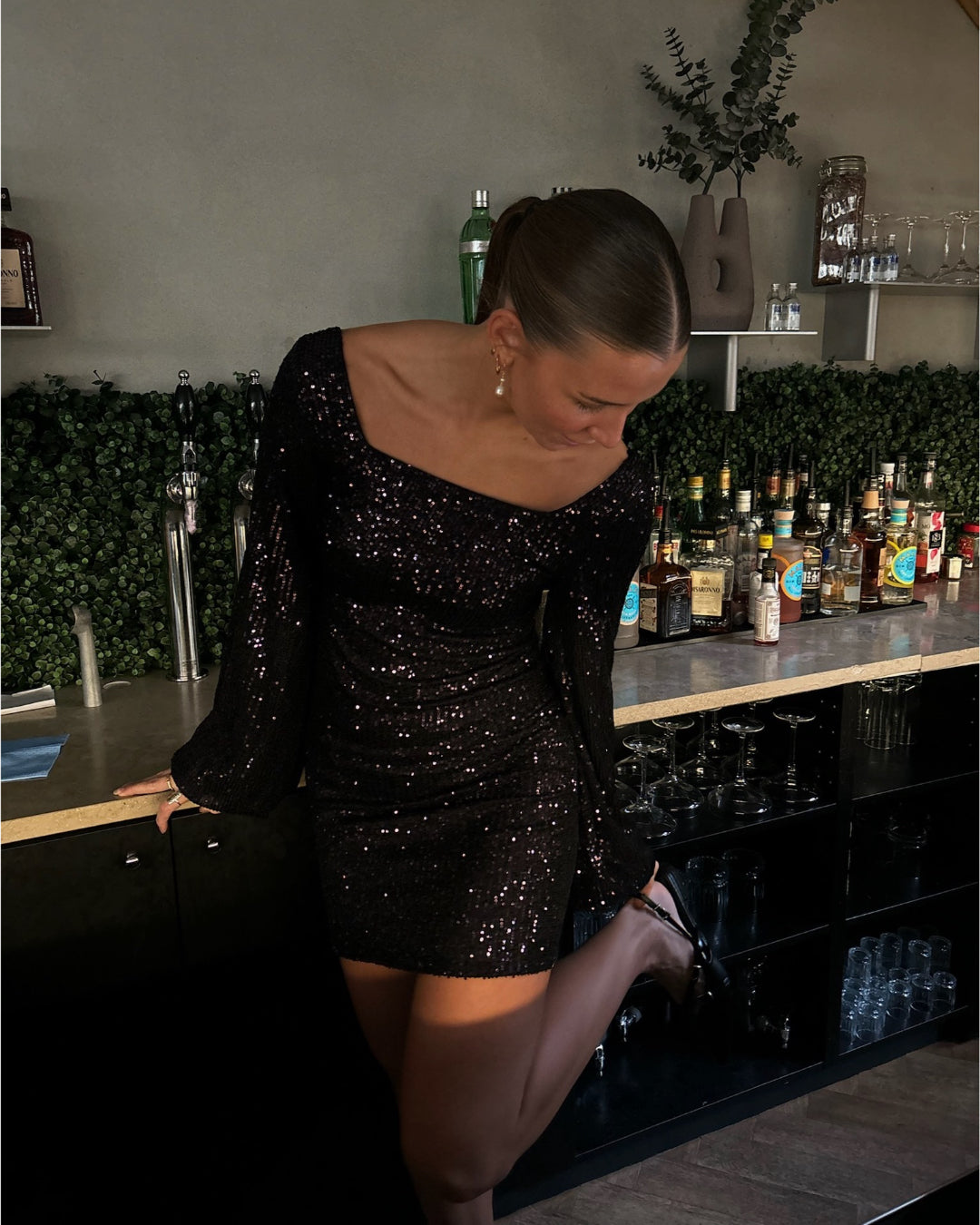 En kvinde iført en glitrende sort kjole læner sig op ad en bar med flasker og glas. Baggrunden har grønne planter og hylder med spiritusflasker.