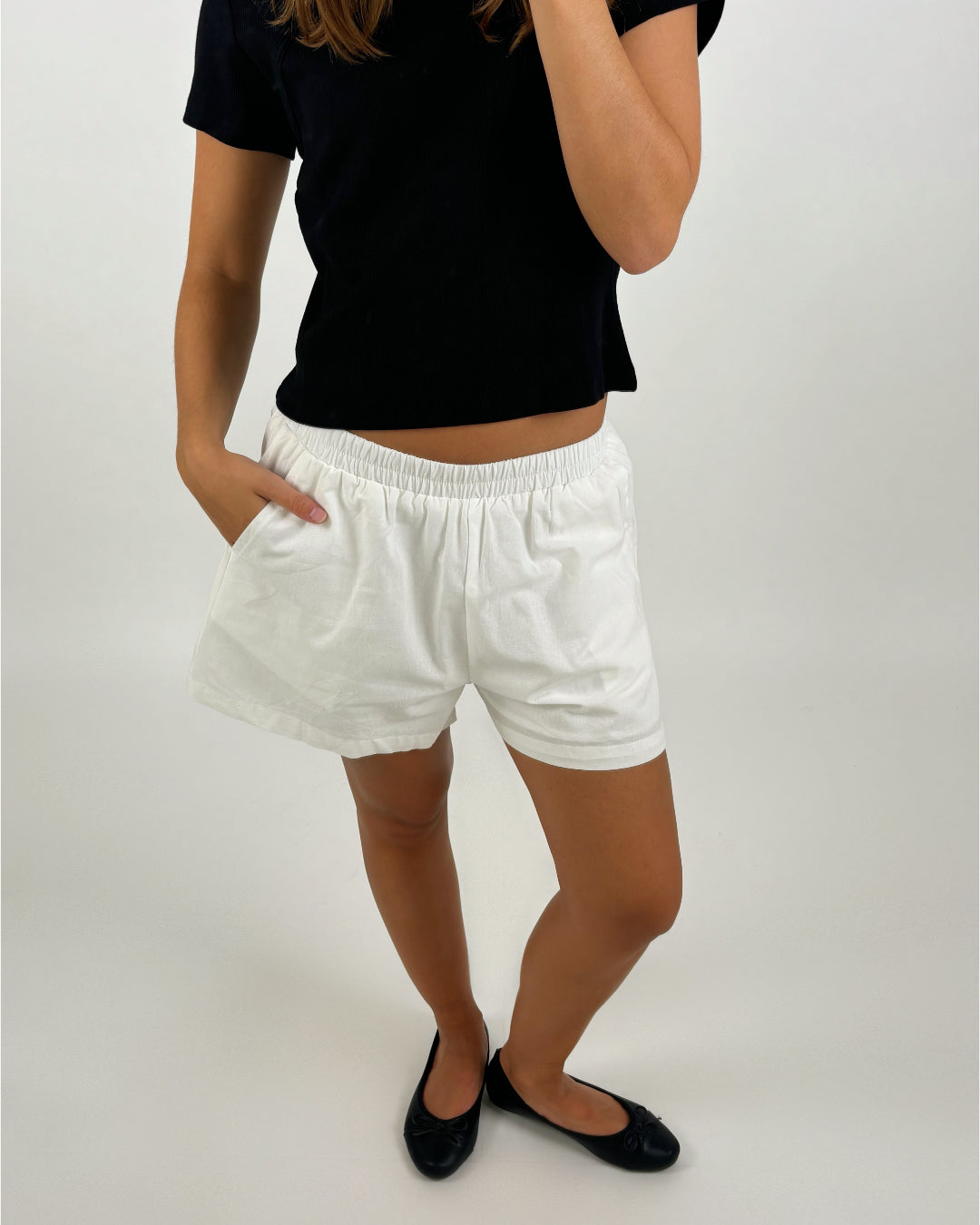 En person iført en sort t-shirt og hvide shorts står med den ene hånd i lommen, i et neutralt rum. Personen har sorte flade sko på.