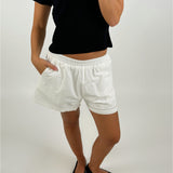 En person iført en sort t-shirt og hvide shorts står med den ene hånd i lommen, i et neutralt rum. Personen har sorte flade sko på.