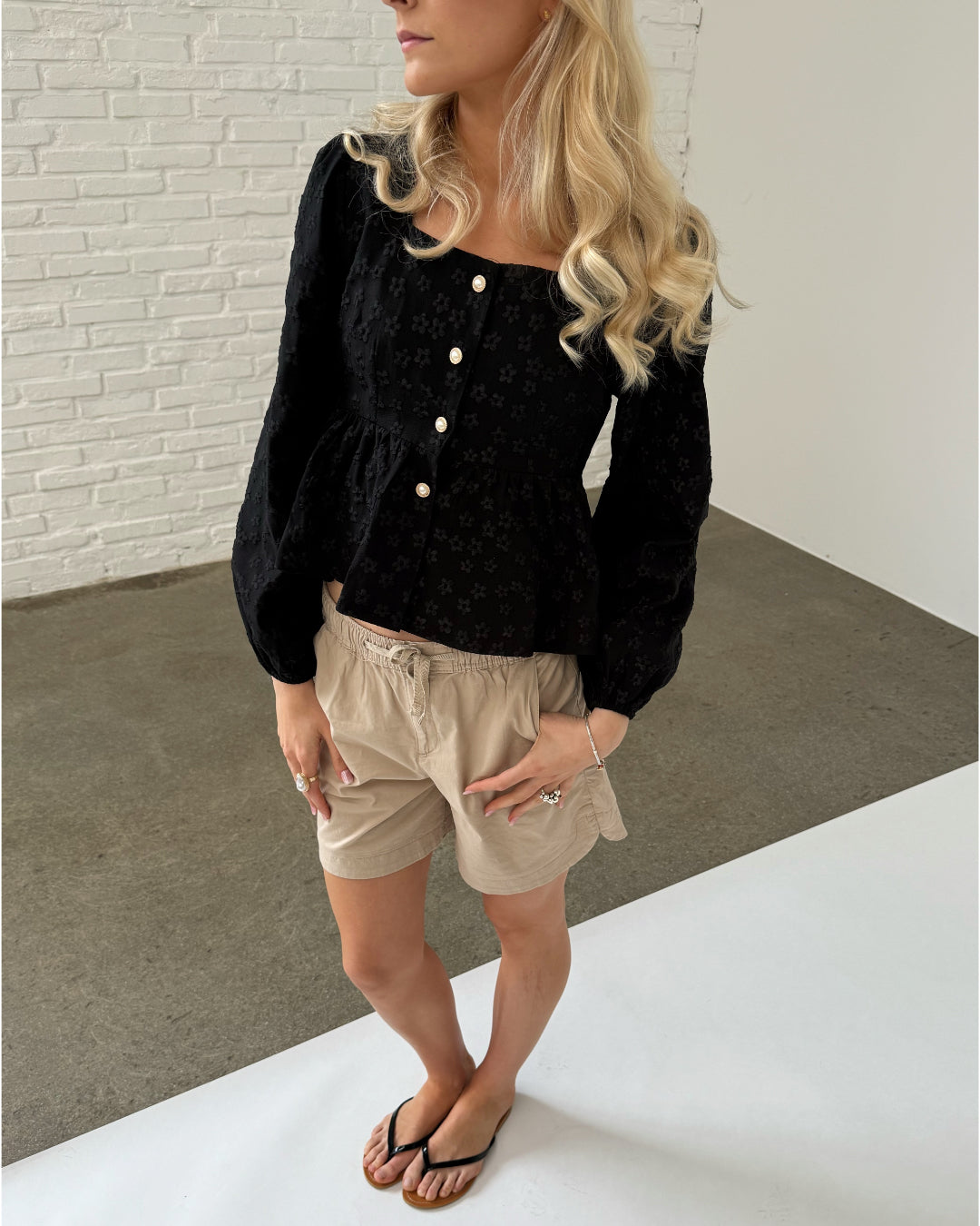 En kvinde med lyst, bølget hår står iført en sort bluse med knapper og beige shorts. Hun har sorte klipklappere på en grå beton- og hvid baggrund.